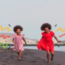 Duas meninas negras, uma de vestido com estampa floral e a outra com vestido vermelho, caminham por uma praia