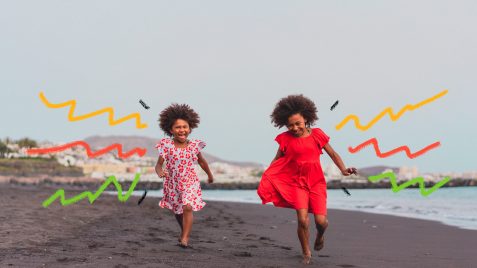Duas meninas negras, uma de vestido com estampa floral e a outra com vestido vermelho, caminham por uma praia