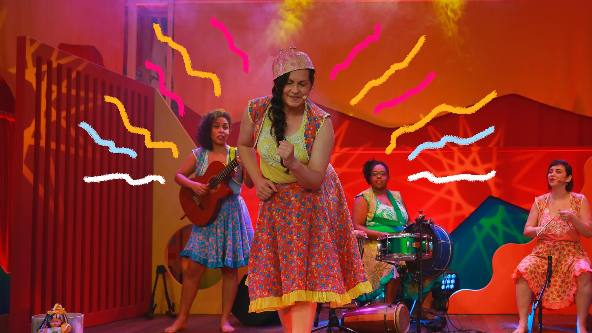 Quatro mulheres estão em um palco, dançando e tocando instrumentos musicais como violão, bateria e triãngulo. Todas elas estão vestindo roupas coloridas e saias de chita.