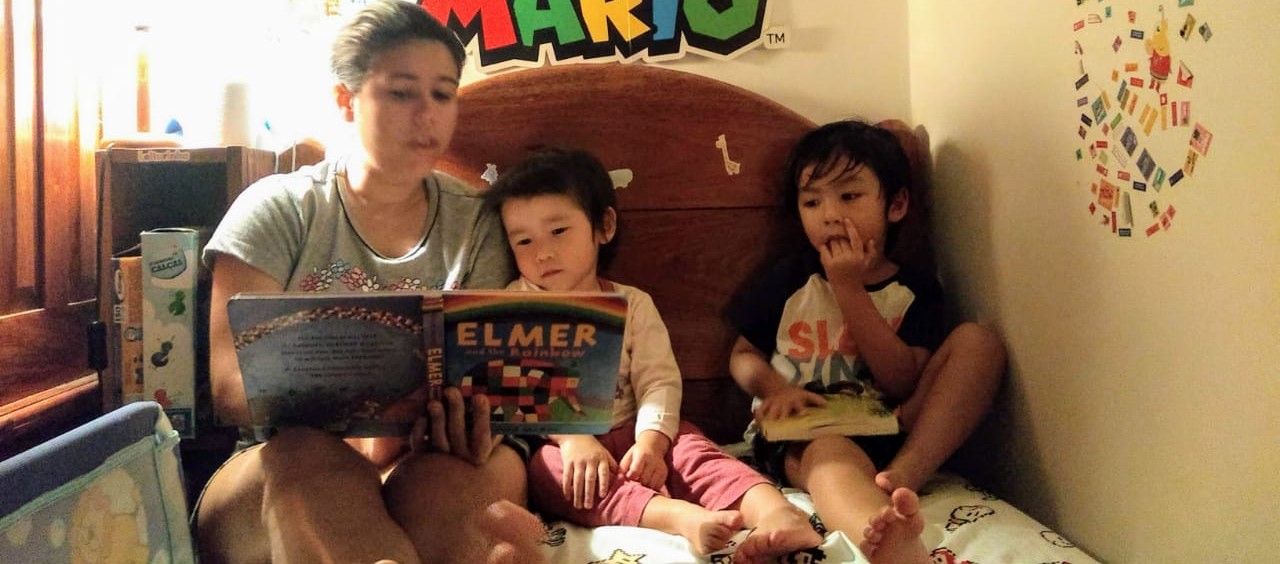 Mulher sentada em uma cama, com duas crianças, lendo um livro