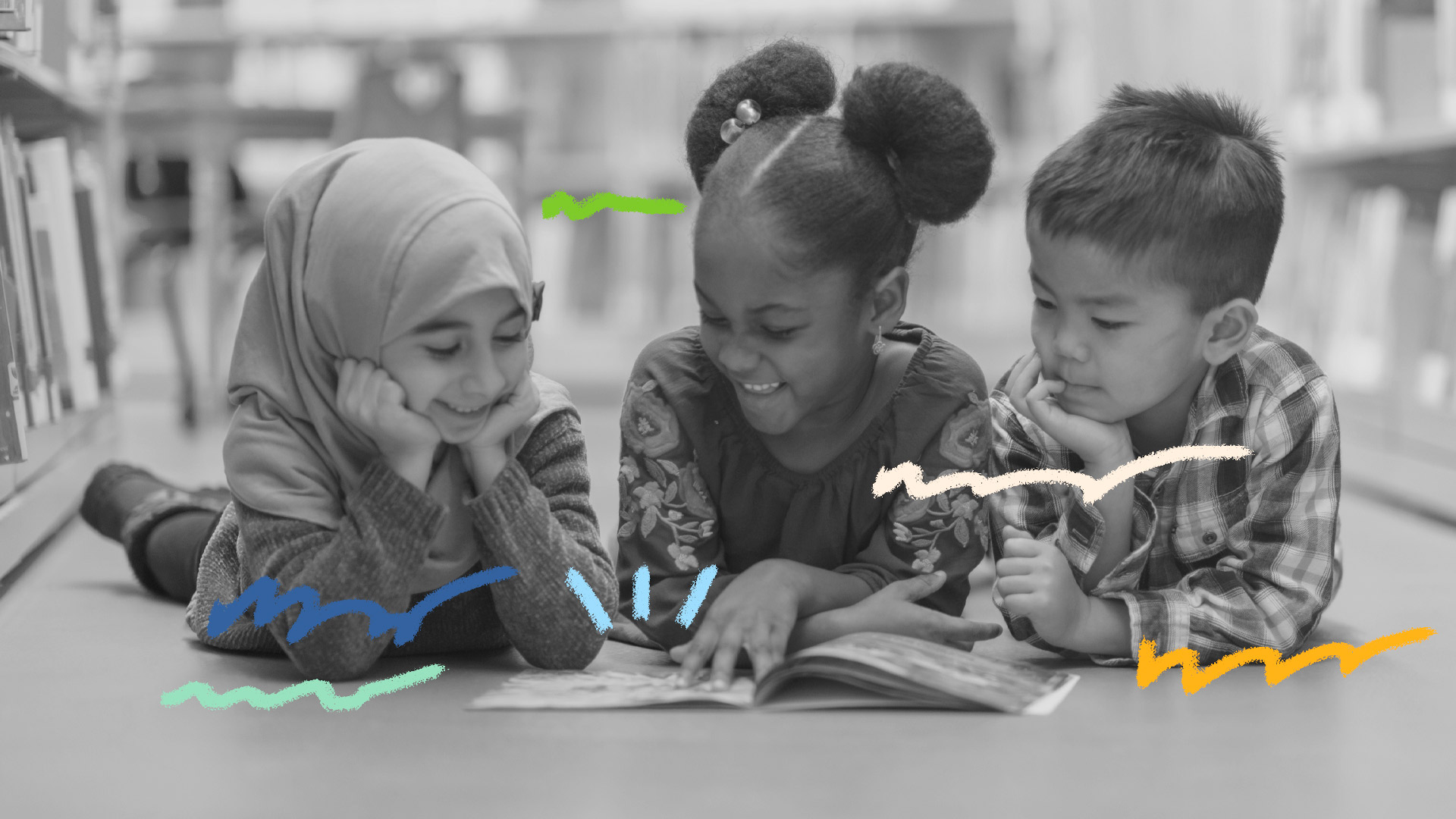 Foto em preto e branco em que três crianças de etnias diferentes leem o mesmo livro deitadas no chão de uma biblioteca
