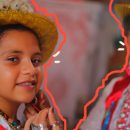 Uma menina sorri, enquanto uma mulher amarra nela um chapéu dourado com plumas brancas. Ambas vestem roupas típicas da festa da Marujada de Bragança