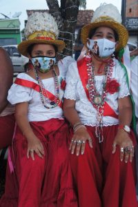 Uma menina e uma senhora, usando máscara no rosto (por conta da Covid-19), vestindo trajes típicos da Festa da Marujada de Bragança: saia longa vermelha, blusa branca, colares e um chapéu dourado com plumas brancas 