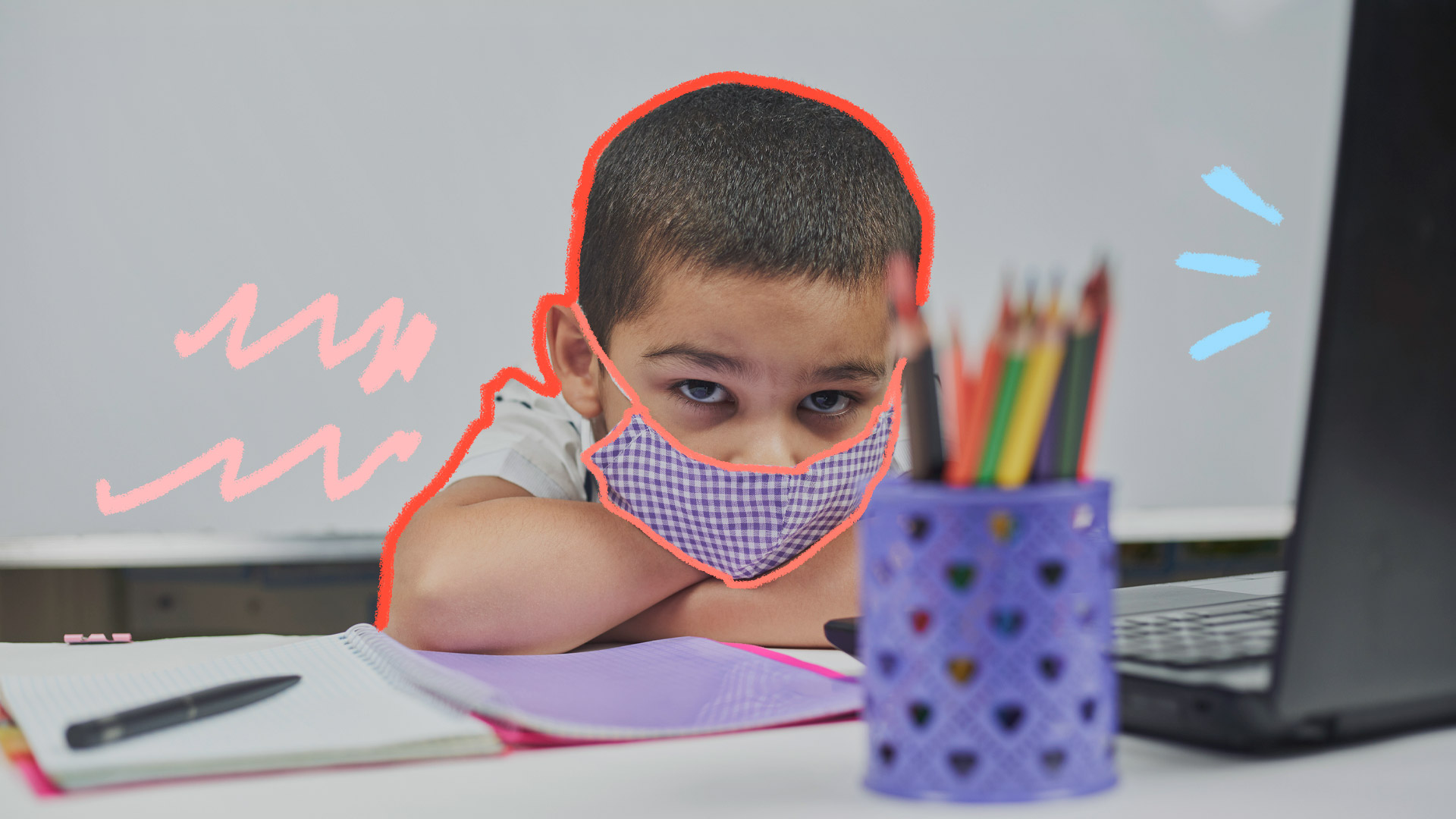 Foto de um menino usando máscara, debruçado em uma mesa, onde estão um caderno, um computador e lápis de cor
