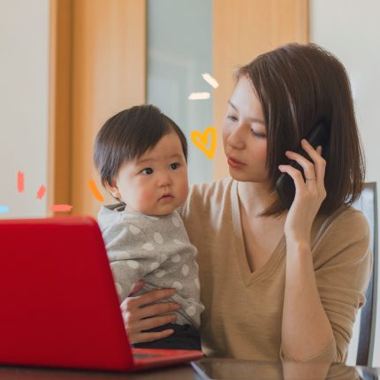 Mães empreendedoras: Uma mulher oriental, com pele branca e cabelos lisos na altura do ombro, está ao telefone, enquanto segura uma criança, também oriental, no colo. Ela está sentada em frente ao computador.