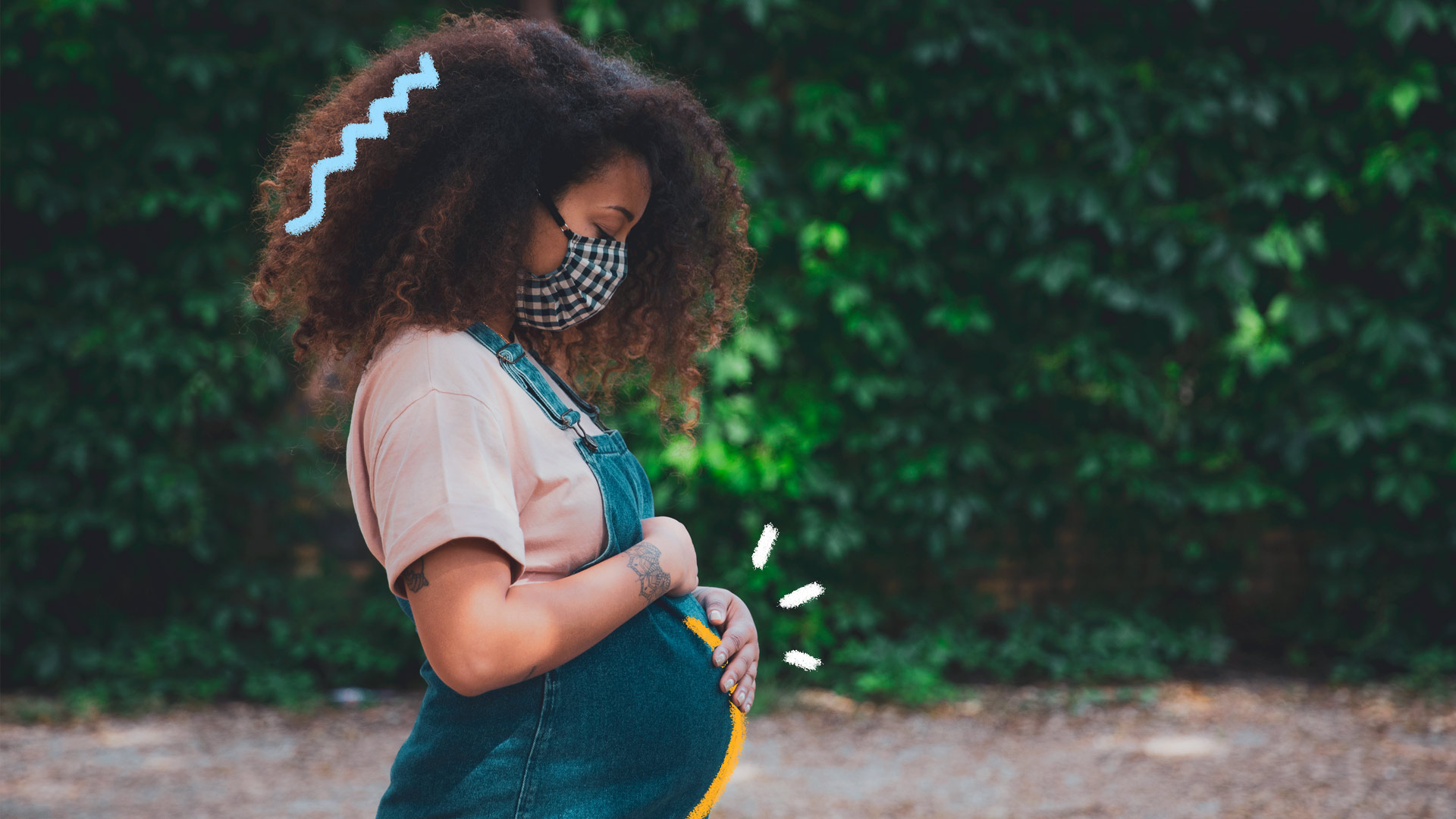 Foto de uma mulher grávida com as mãos na barriga. Ela usa jardineira jeans e camiseta branca. Está posicionada de perfil em frente à uma parede de folhagens