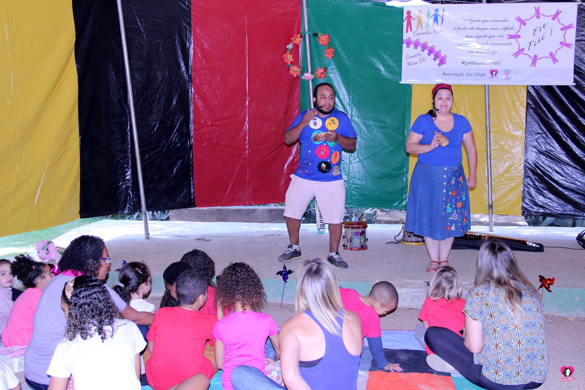 Um casal vestido com roupas azuis está num palco com lona colorida atrás, se apresentando a um grupo de crianças