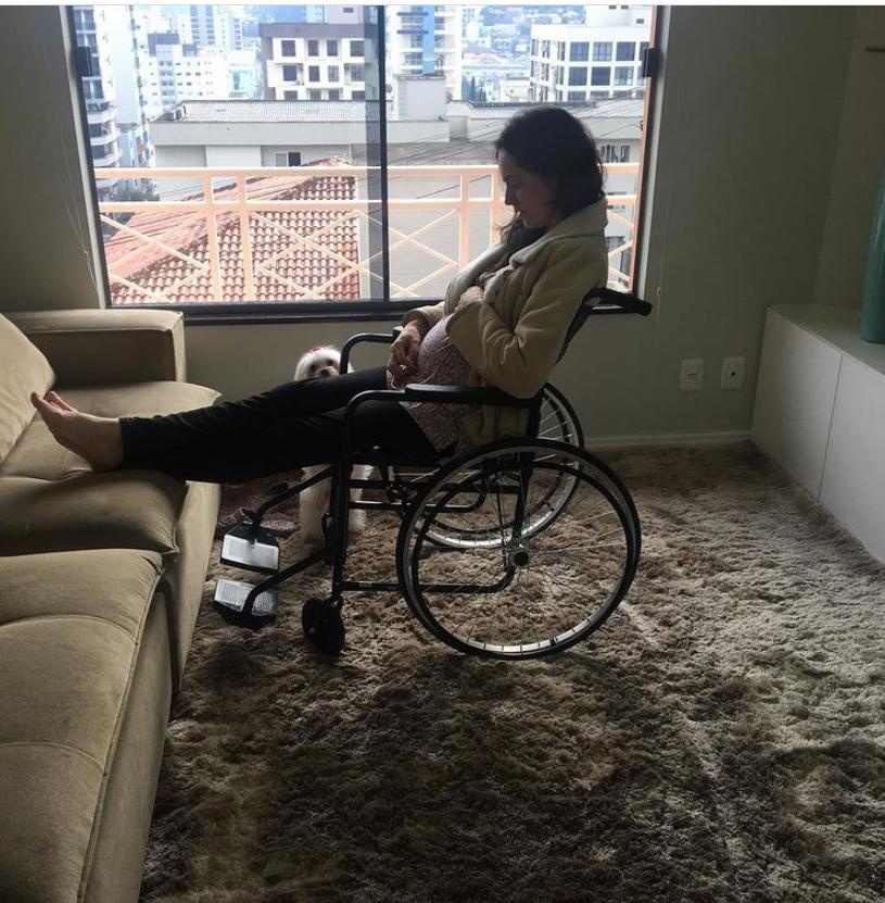 Uma mulher grávida na sala de sua casa em um apartamento, em uma cadeira de rodas e com os pés esticados no sofá