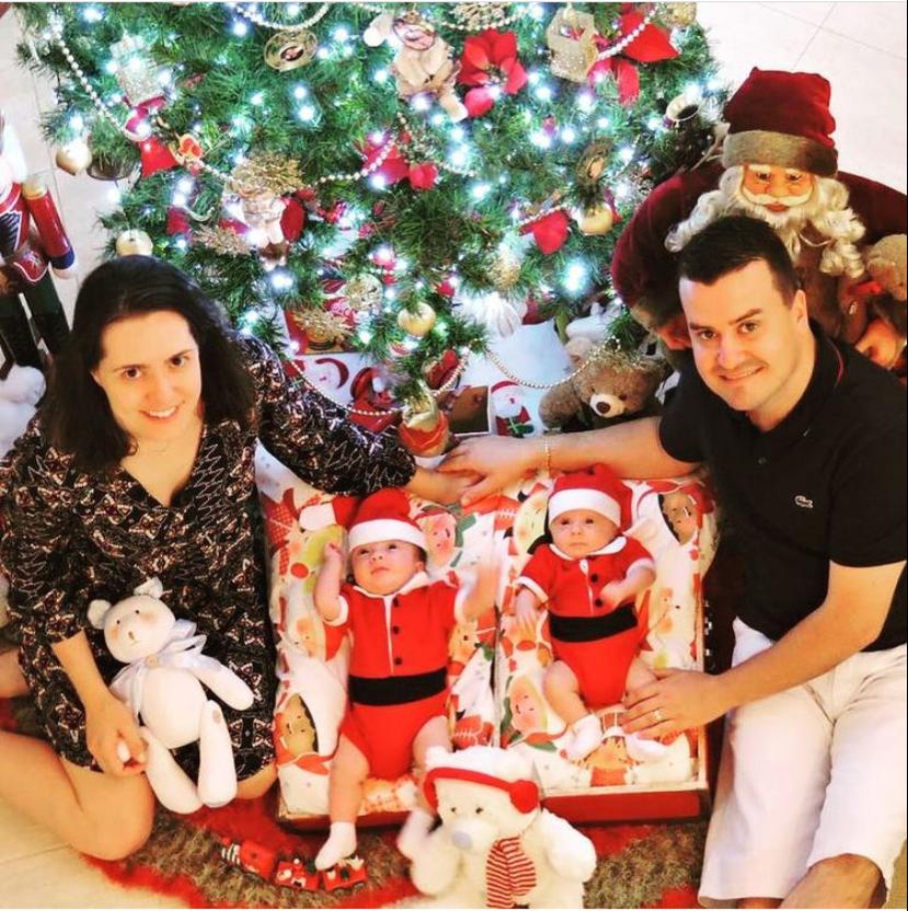 Casal sentado com dois bebês vestidos de Papai Noel em frente a uma árvore de Natal