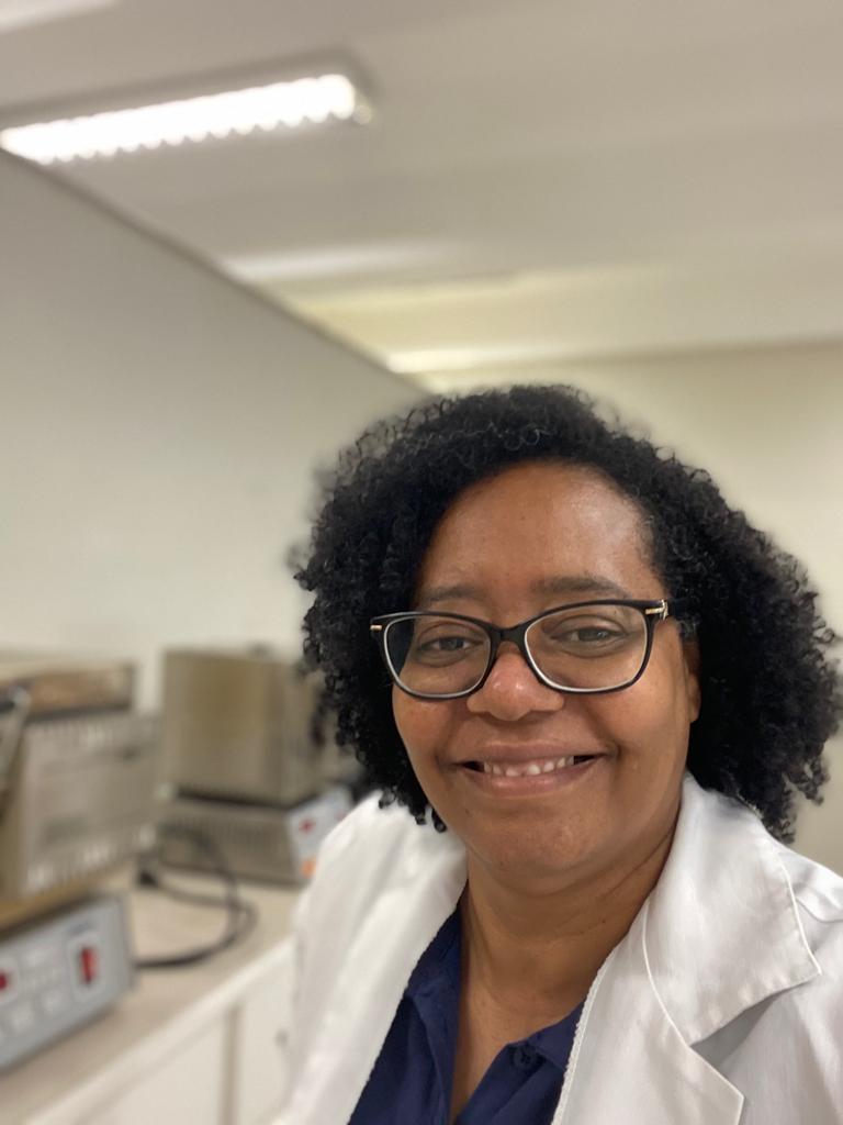 mães na ciência: mulher negra de cabelos crespos e óculos, vestindo um jaleco branco, em um laboratório com computadores ao fundo