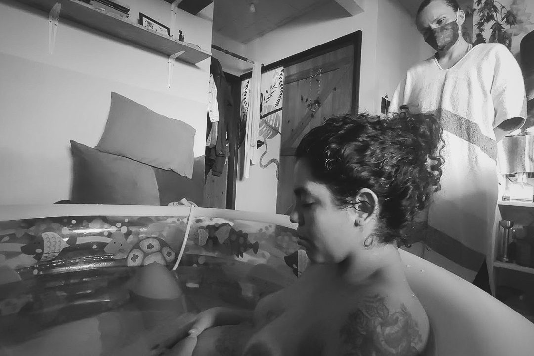 Foto em preto e branco de uma mulher dentro de uma banheira na hora do parto, em casa. Ao lado, a doula acompanhando-a.