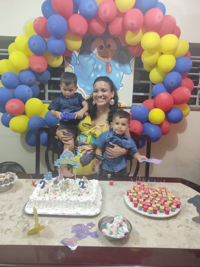 Uma mulher com dois filhos no colo, em frente a uma mesa com bolo de aniversário e doces, e um arco de balões coloridos ao fundo