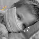 Foto em preto e branco de uma criança, com máscara, deitada em um leito, com olhar triste