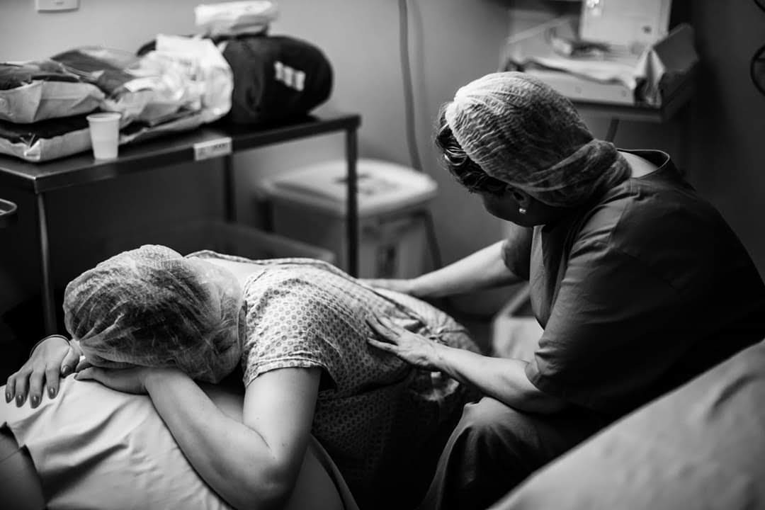 Foto em preto e branco de uma mulher deitada de bruços em uma bola, e a doula fazendo uma massagem em suas costas - ambas estão vestindo toucas e aventais hospitalaraes 