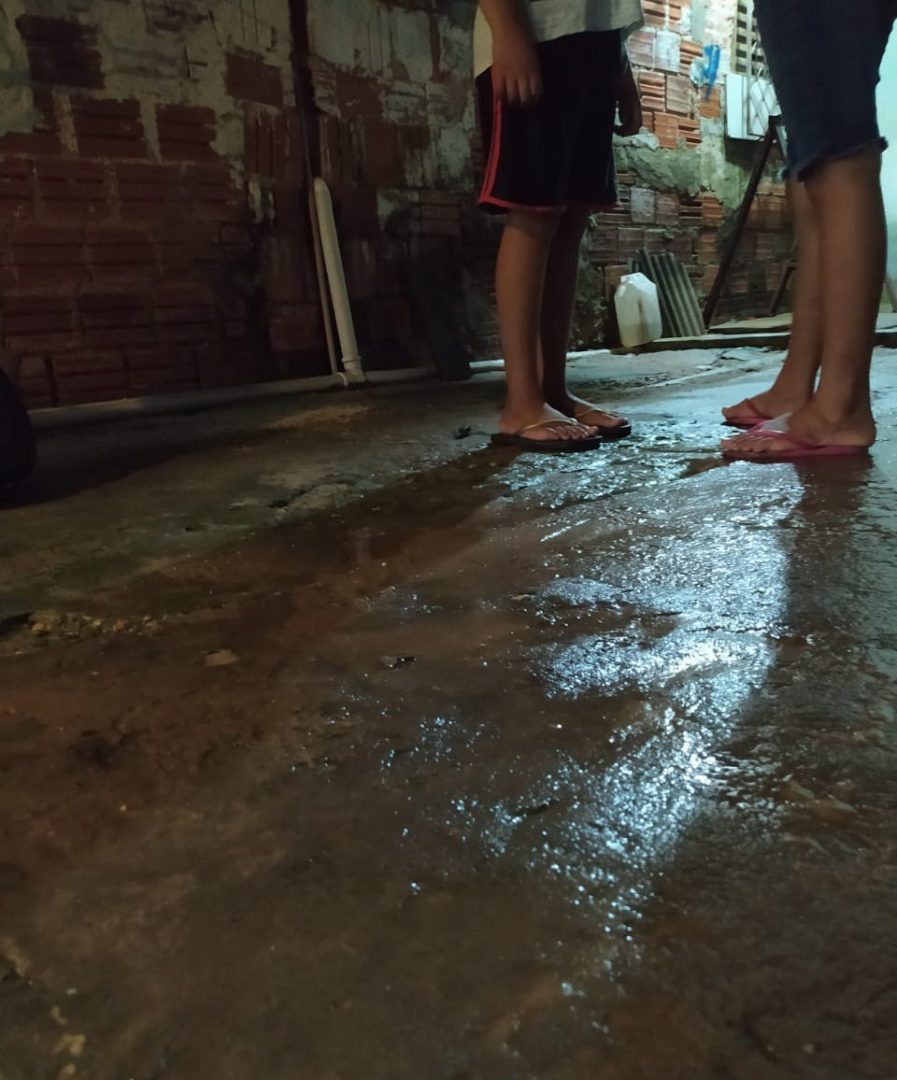 Foto mostra os pés de duas crianças, usando chinelos, em um chão molhado de cimento