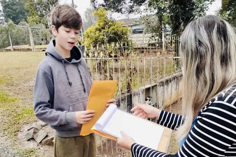 Uma professora com uma prancheta na mão entregando um envelope a um menino, que vive num ambiente rural