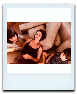 Foto tirada de cima para baixo, mostrando uma mulher de cabelos curtos e negros, deitada em um sofá, com os dois filhos ao lado (um menino do lado direito e uma menina do lado esquerdo)