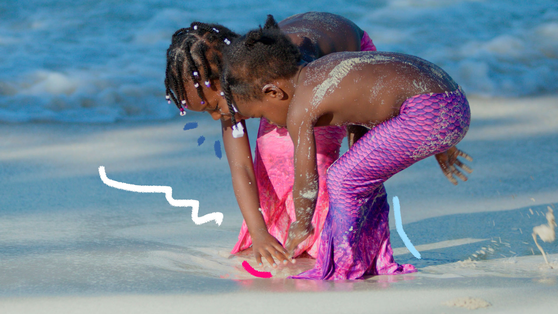 Duas meninas com caudas de sereia brincam com areia em uma praia