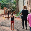 Uma criança está de braços dados com duas mulheres, lado a lado, andando por um terreno de chão batido, em um lugar de moradias irregulares