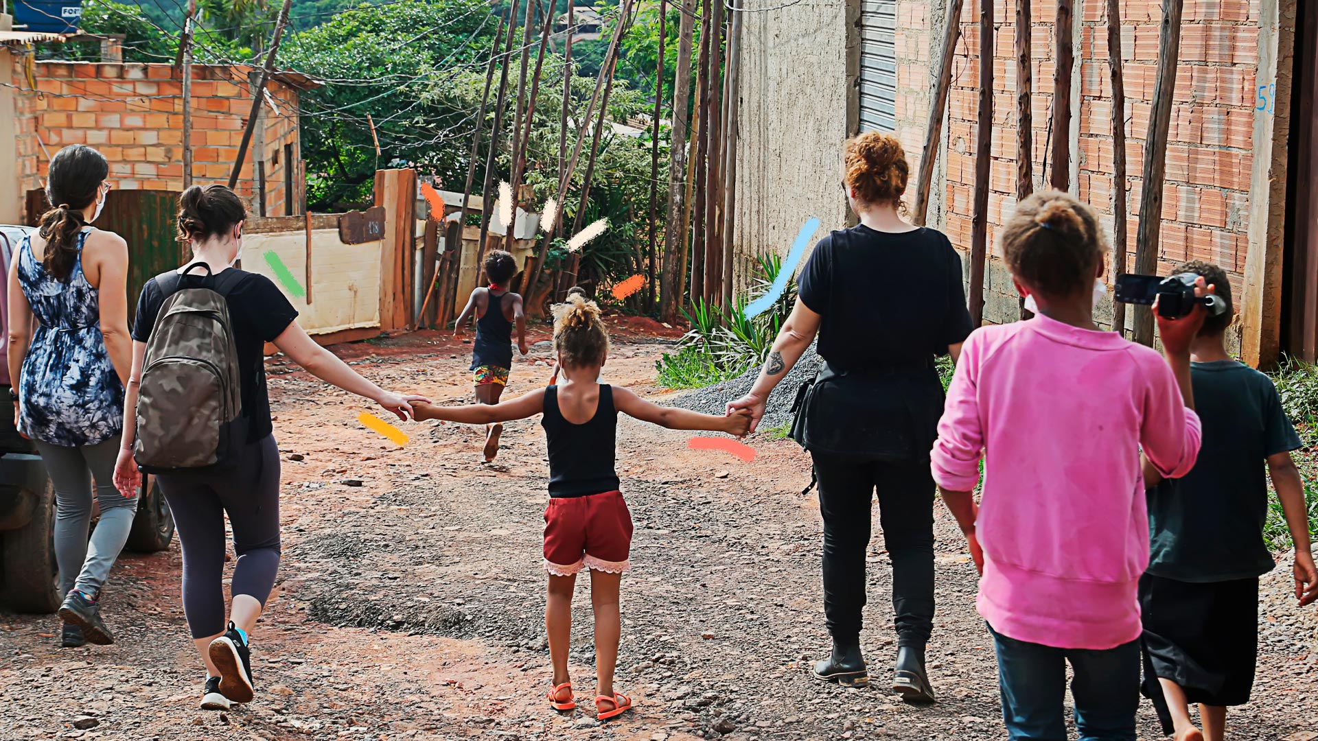 Uma criança está de braços dados com duas mulheres, lado a lado, andando por um terreno de chão batido, em um lugar de moradias irregulares