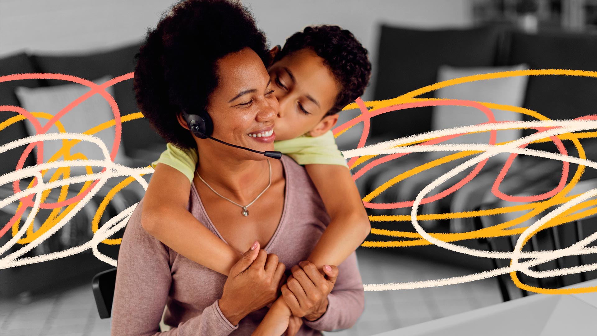 Empresas family-friendly: uma mãe usando um fone de ouvido com microfone, sentada em frente a um computador, recebe um abraço e um beijo de seu filho, que a abraça por trás.