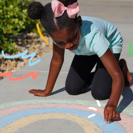 Uma menina negra rabisca com giz um arco-íris no chão.
