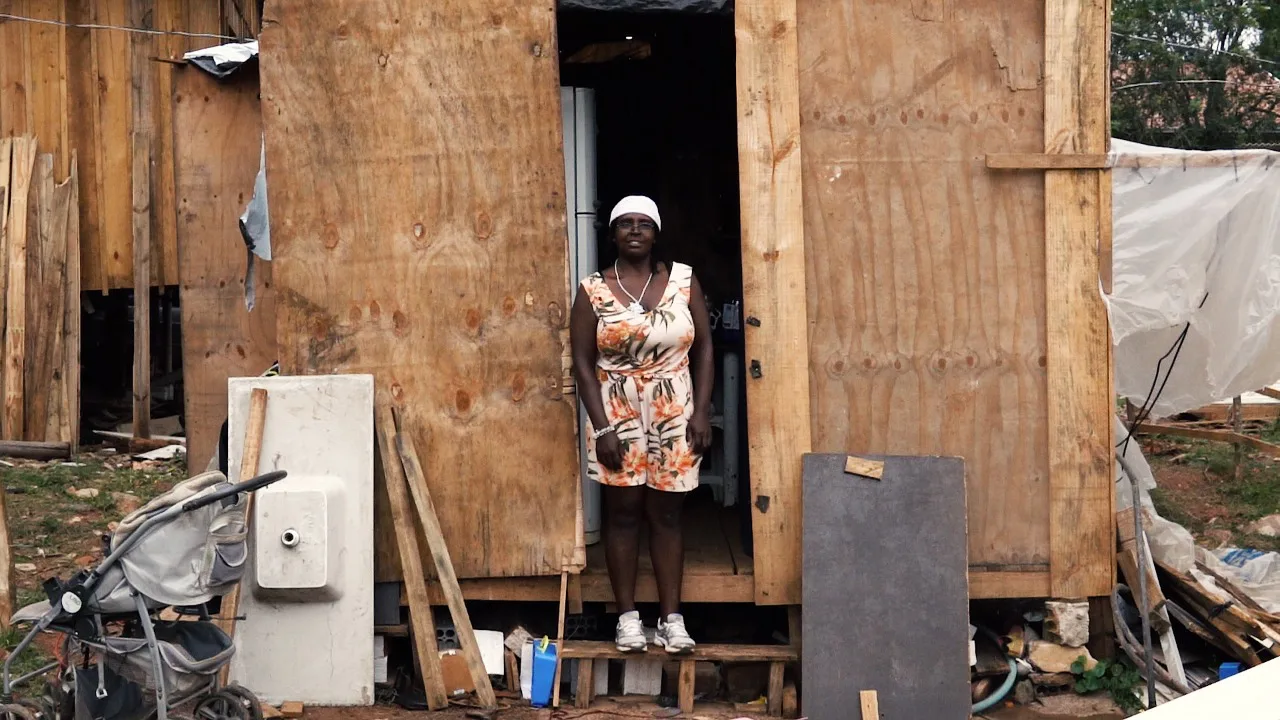 Fome e pandemia: mulher de pele negra está com um lenço branco na cabeça, vestido florido, em frente a uma porta de uma casa feita de tábuas, em uma favela
