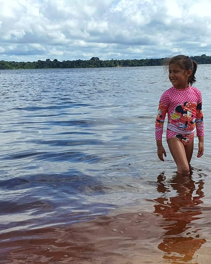 Crianças da Amazônia: Foto de uma menina vestindo camisa manga longa e shorts, andando em um rio com água até o joelho