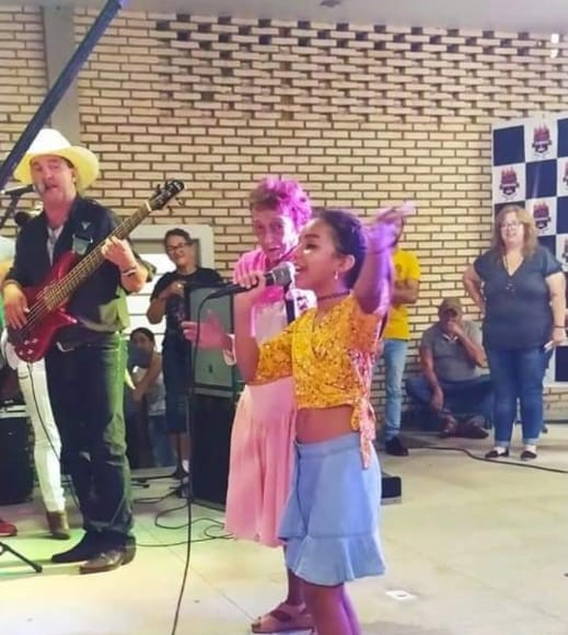 Música sertaneja e infância: Uma menina (Rafaela), com microfone na mão, está se apresentando a um público pequeno, com uma senhora ao lado cantando (Delinha)