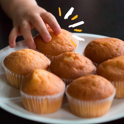 Foto de um prato branco com bolinho de banana (7 bolinhos em forminhas separadas) e uma mão de uma criança pegando um deles