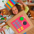 Brincadeiras para reciclagem: Uma menina, de pele negra e cabelos crespos, está dentro de uma caixa de papel e usando um chapéu feito de caixa e papel colorido