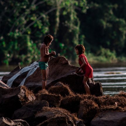 Duas crianças yanomami, um menino e uma menina, estão em cima de uma pedra, na frente de um rio