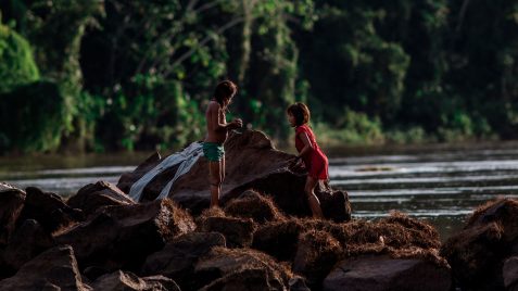 Duas crianças yanomami, um menino e uma menina, estão em cima de uma pedra, na frente de um rio
