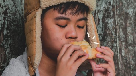 Um menino, usando uma touca, está de olhos fechados com um sanduíche na mão