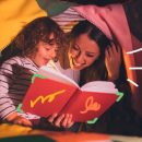 Foto de uma mãe lendo para uma criança. A dupla está com o livro aberto embaixo de uma cabaninha improvisada com lençóis em rosa e amarelo