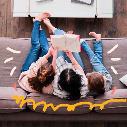 Mães na quarentena: De cima, a foto registra uma mãe lendo um livro para seus dois filhos. Cada criança está sentada de um lado do sofá e a mulher no meio segurando o livro.