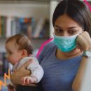 Famílias na pandemia: Uma mãe segura seu bebê no colo. Ela usa máscara. Ao fundo, há uma estante de livros