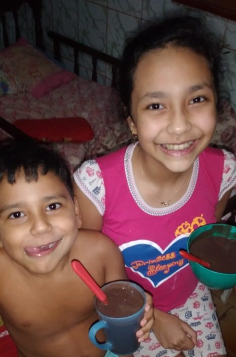 Um menino mais novo e uma menina, ambos de pele de cor parda, estão sorrindo e segurando uma tigela de açaí da amazônia