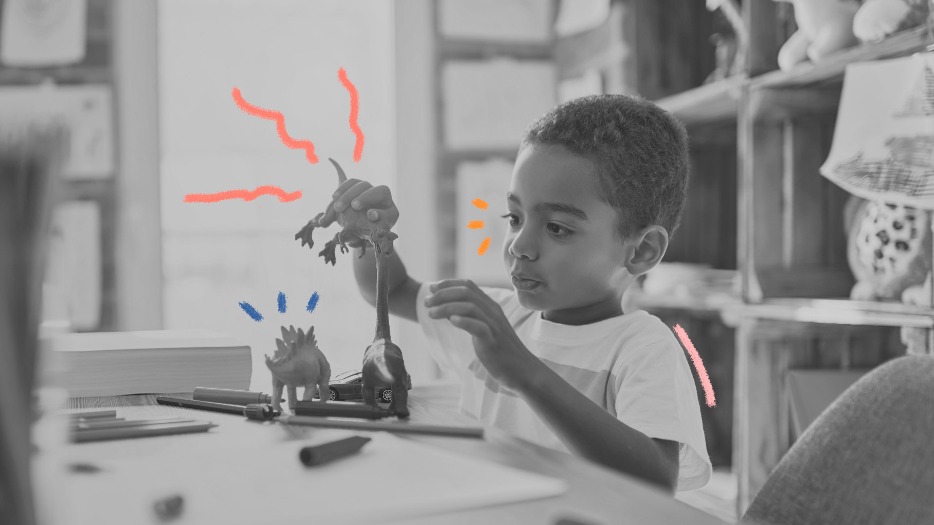 Foto em preto e branco, de um menino negro vestindo camiseta branca, brincando com dinossauros de brinquedo em cima de uma mesa