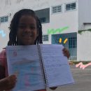 Foto de uma menina negra, de cabelos encaracolados, em frente à Escola-Creche 11 de Dezembro, com um caderno desenhado nas mãos