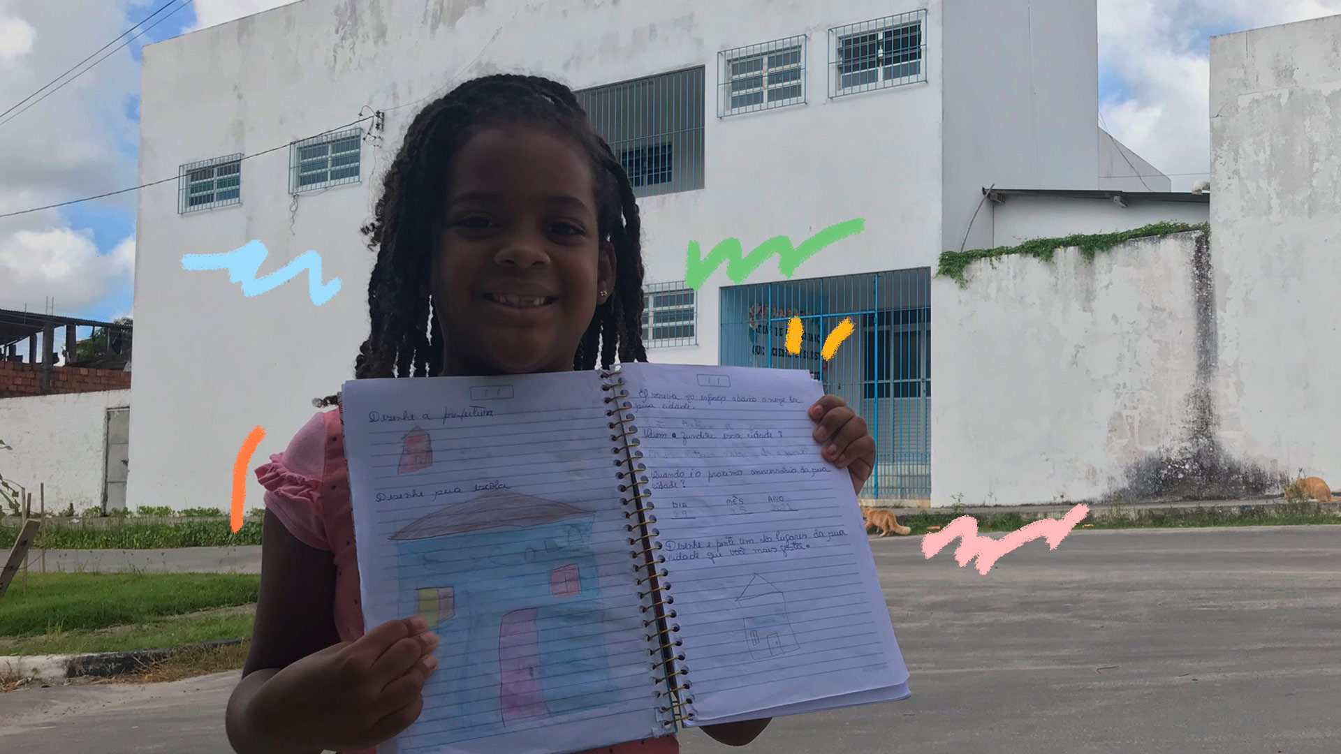 Foto de uma menina negra, de cabelos encaracolados, em frente à Escola-Creche 11 de Dezembro, com um caderno desenhado nas mãos
