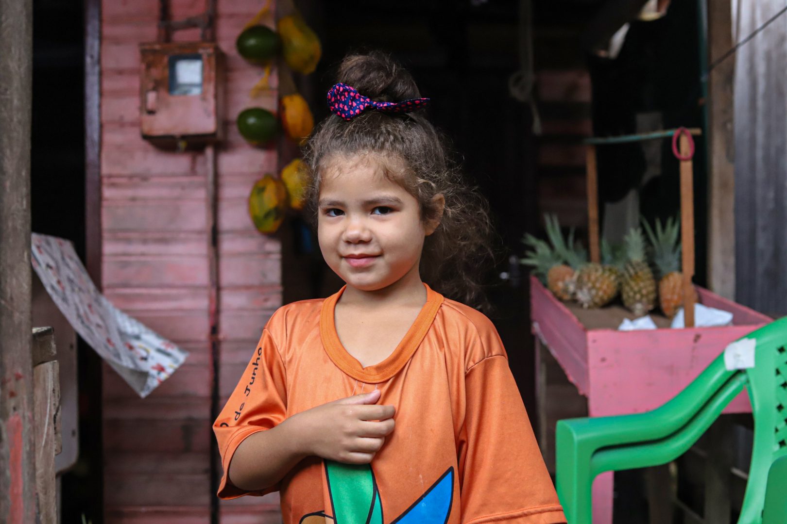Uma menina de cabelos cacheados e castanho claro, vestindo uma camiseta laranja, posa para a foto, em frente a um mercadinho de madeira, onde estão expostas algumas frutas.