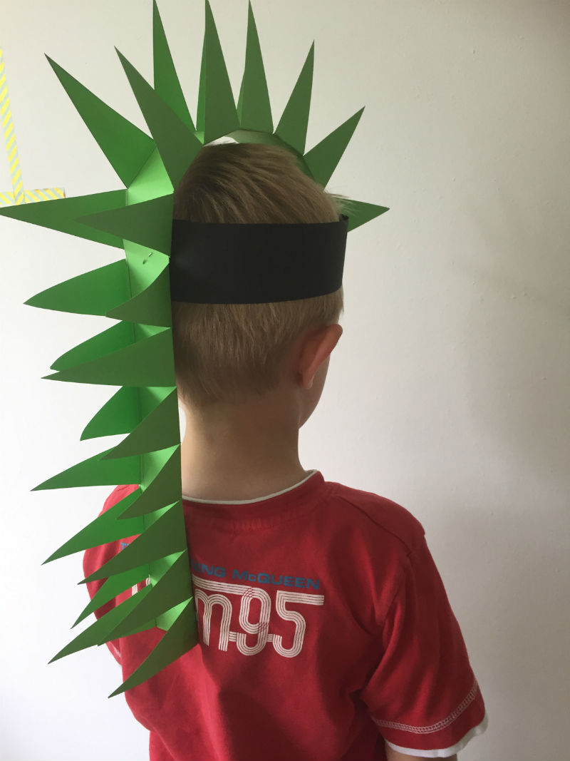 Brincadeira de dinossauro: foto de um menino, de costas, com um chapéu de cartolina verde que imita a coluna/espigões do dinossauro, que vai até o meio das suas costas