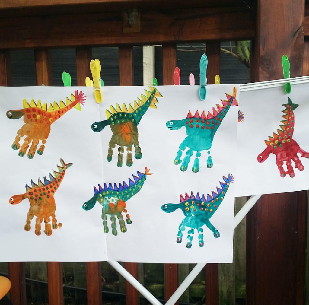 Brincadeiras de dinossauros: folhas de papel em um varal presas com grampo colorido, onde há vários dinossauros pintados com tinta guache