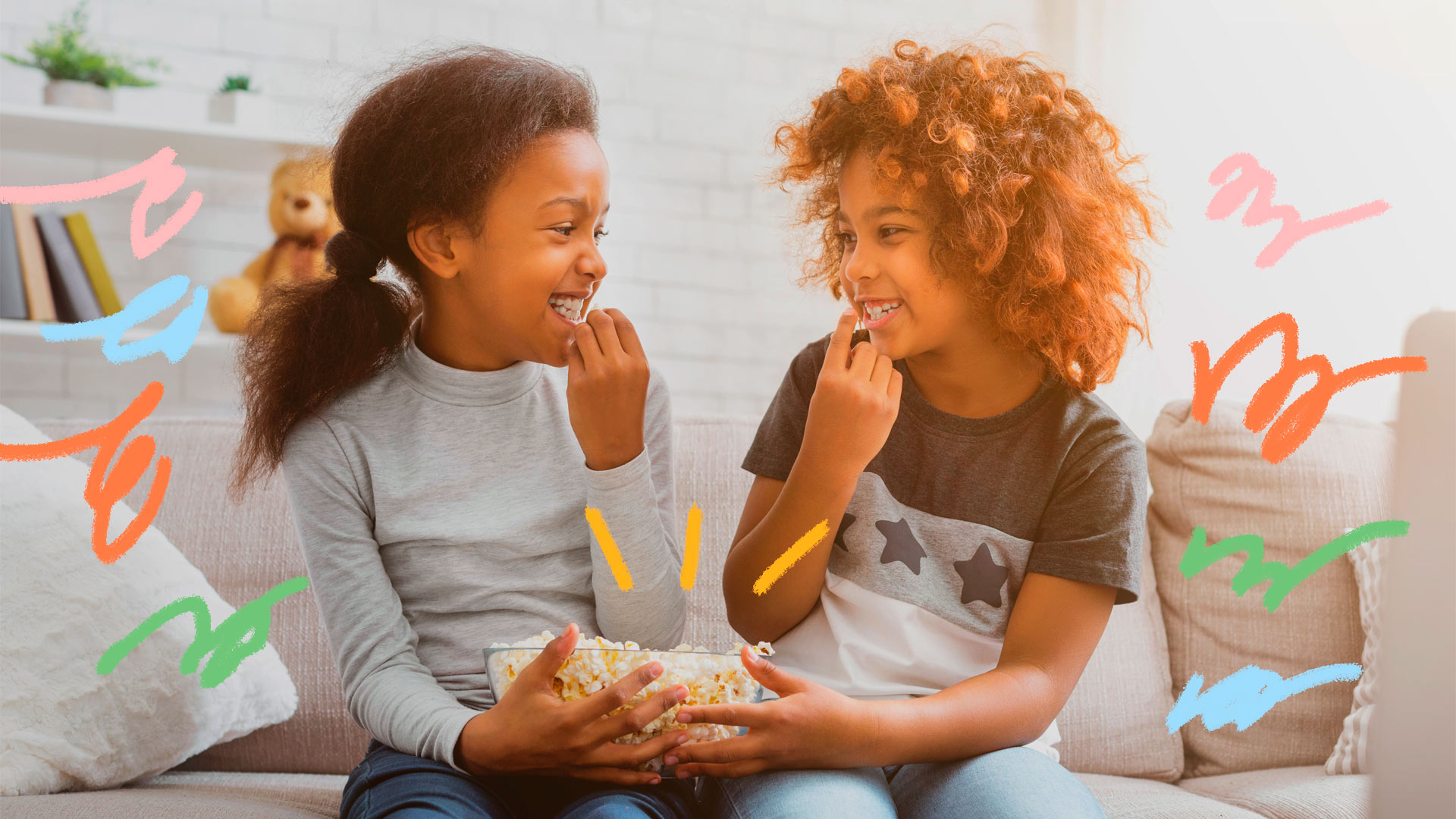 Na foto, duas meninas negras sorriem enquanto se olham e dividem uma pote com pipoca