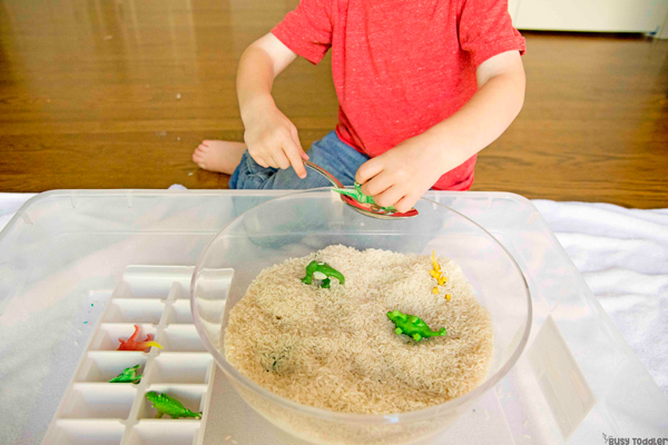Brincadeiras de dinossauros: uma criança (não aparece seu rosto), está enterrando pequenos brinquedos de dinossauros em uma tigela transparente cheia de areia