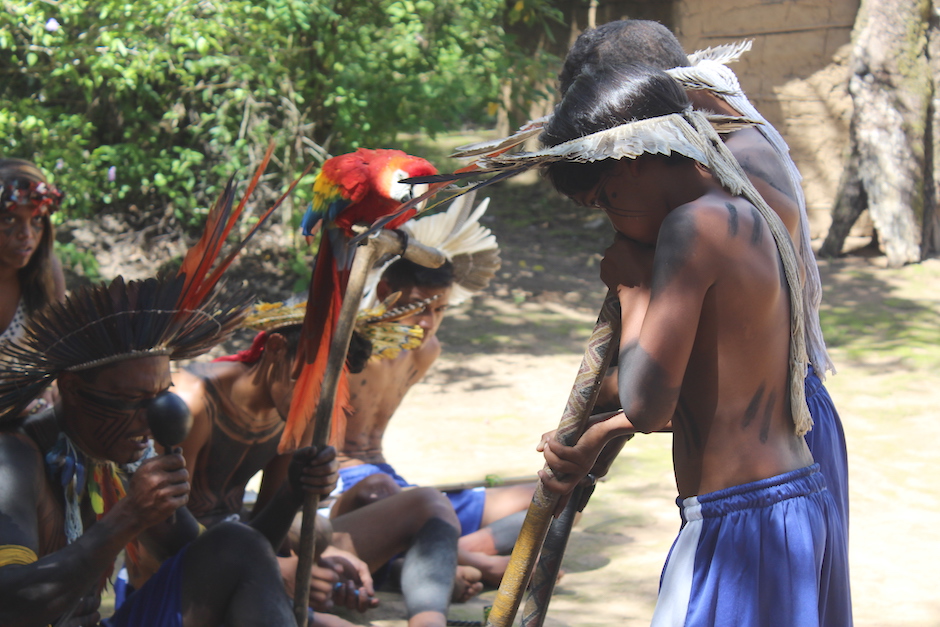 De pé, uma criança Xukuru-Kariri está tocando uma flauta grande de madeira, usando um cocar. Sentados, estão os indígenas mais velhos