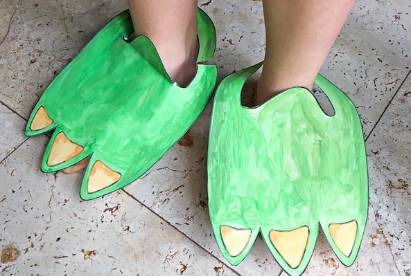 Brincadeiras de dinossauros: uma criança de pele clara usa um 'sapato de cartolina' com formato de pé de dinossauro, de cor verde e unhas pontudas amarelas