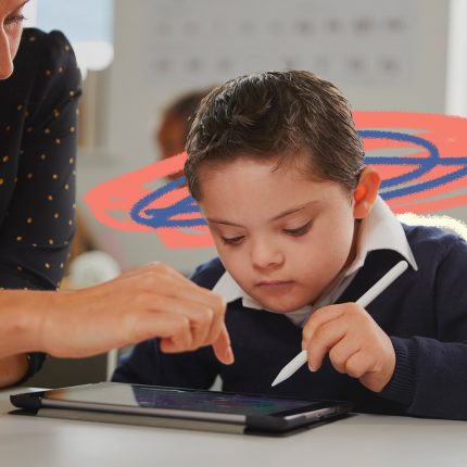 Na foto, um menino de pele clara utiliza um tablet com auxílio de uma caneta, com orientação de sua professora