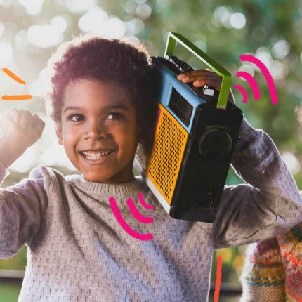 Duas crianças, ambas negras de cabelos curtos e crespos, estão sorrindo. Uma delas está carregando um radinho de pilha, antigo, e parece vibrar com o som.